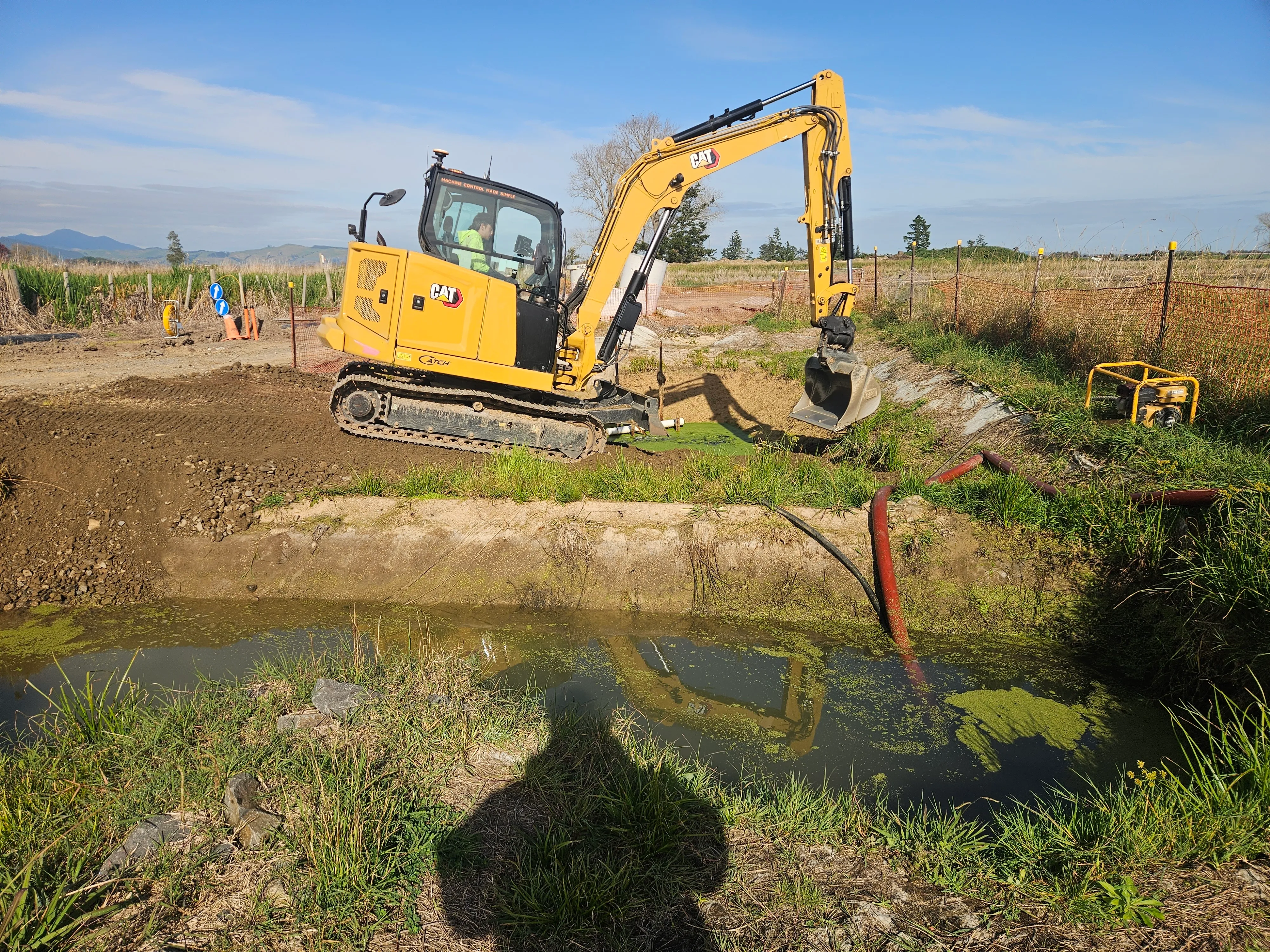 Excavator cleaning a farm drain beside a green paddock